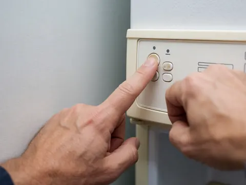 Main ajustant le contrôle de température d'un réfrigérateur avec un panneau comportant des boutons et des détails en plastique sur un fond de mur gris clair.