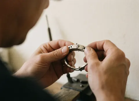Mains d'un technicien ajustant avec précision une fermeture de montre en métal brillant sur un fond de atelier flou aux murs clairs.