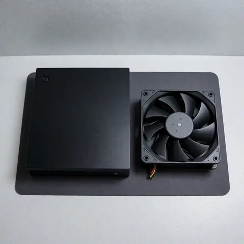 Console de jeux vidéo noire posée sur une surface sombre, accompagnée d'un ventilateur de rechange neuf, évoquant un service de réparation et de maintenance matérielle.
