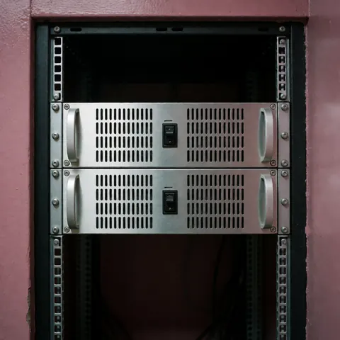 Deux amplificateurs professionnels métalliques avec ventilation, installés dans un rack de 19 pouces sur fond rose, éclairage froid et technique pour événements.