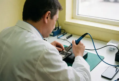 Un technicien répare un téléphone avec un fer à souder sur une table devant un mur jaune en lumière naturelle.