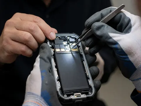 Un technicien en gants nettoie les contacts internes d'un smartphone robuste à l'aide de pinces, sur un fond sombre.