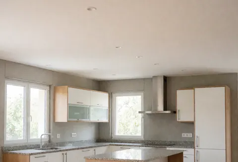 Cuisine ouverte moderne dotée d'armoires blanches, d'un plan de travail en granit, de murs en béton et de fenêtres laissant entrer la lumière naturelle.
