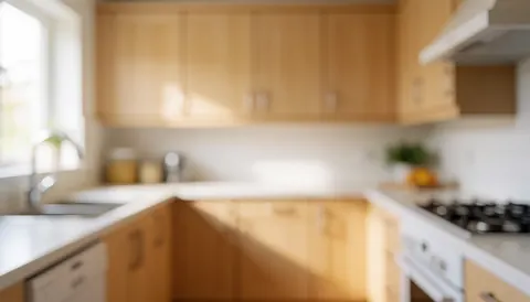 Intérieur de cuisine résidentielle avec placards en bois clair, surface blanche lisse et appareils ménagers, arrière-plan flou pour texte, éclairage naturel chaleureux.