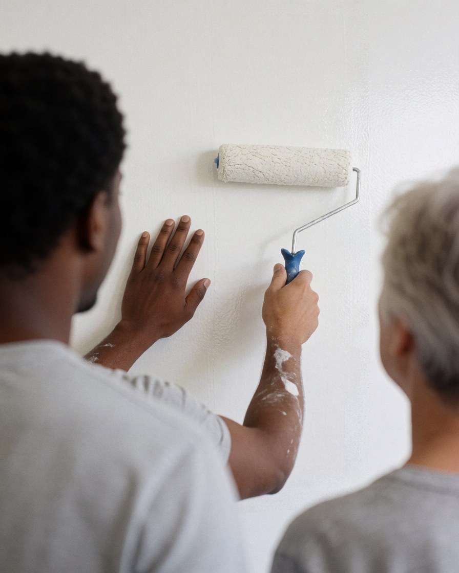 Un ouvrier peint une paroi blanche à l'aide d'un rouleau tandis qu'une autre personne observe la scène depuis l'arrière-plan.
