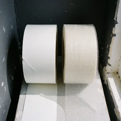Deux rouleaux de ruban adhésif, l'un en papier blanc et l'autre en treillis de verre, posés sur une surface de réparation avec des murs peints en gris anthracite.