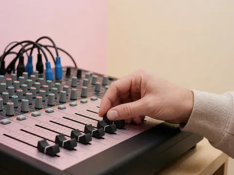 Gros plan d'une main réglant un slider sur un équipement de sonorisation professionnel avec des câbles visibles sur un fond aux tons pastel.