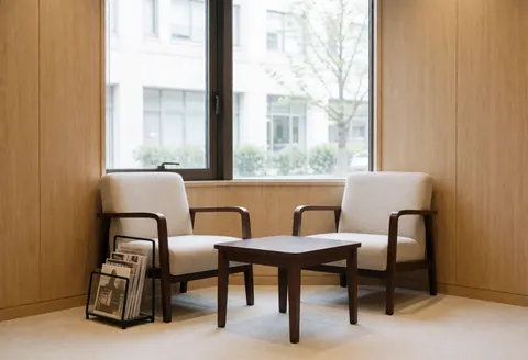 Salle d'attente équipée de deux fauteuils rembourrés, d'une table basse et d'une étagère à magazines près d'une fenêtre laissant entrer la lumière naturelle.