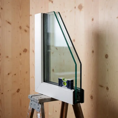 Fenêtre à isolation thermique avec double vitrage, niveau à bulle et cadre blanc posés sur une échelle en bois, devant des murs en panneaux clairs et un éclairage professionnel.