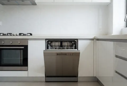 Cuisine moderne avec lave-vaisselle intégré ouvert montrant des étagères vides, murs carrelés blancs et appareils en acier inoxydable.