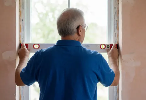 Dos et mains d'un installateur de fenêtres isolantes utilisant un niveau pour mesurer une nouvelle fenêtre dans une pièce aux murs en plâtre et à la lumière naturelle.