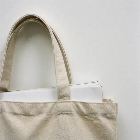 Sac tote beige en toile contenant un cahier blanc aux pages blanches, coutures apparentes sur fond blanc uni pour mise en scène de cours en ligne.