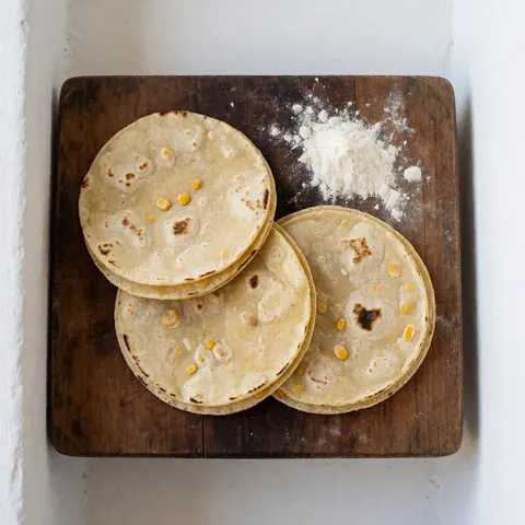 Des tortillas de maïs fraîchement préparées avec des grains visibles posées sur une planche en bois sombre parsemée de farine, éclairées par la lumière naturelle.