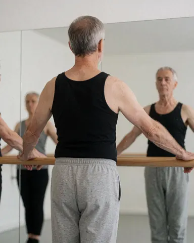 Un homme senior vu de dos dans un studio de danse, tenant une barre en bois. Un miroir reflète sa posture sur un mur blanc. Il porte des vêtements de sport gris et noirs.