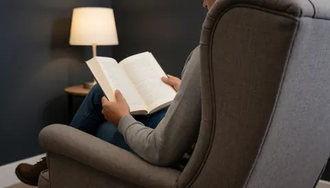 Une personne adulte lit un livre ouvert dans un fauteuil gris avec une lampe allumée à côté, créant une ambiance calme et relaxante.
