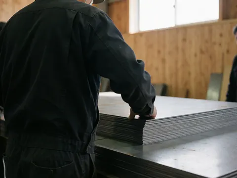 Un ouvrier en atelier métallurgique examine attentivement une pile de tôles découpées, baigné de lumière naturelle dans un environnement aux murs en bois.