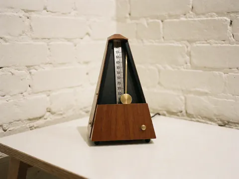 Métronome traditionnel en bois et laiton posé sur une table blanche devant un mur de briques blanches, évoquant précision et musique.