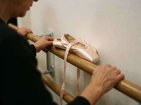 Des chaussures de ballet roses posées sur une barre de bois poli. Des mains d'adulte ajustent la barre dans un studio aux murs blancs et à l'éclairage chaleureux.