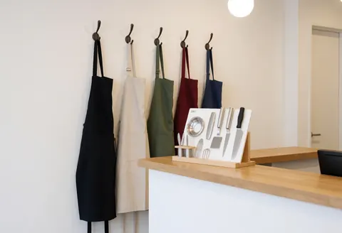 Des tabliers de cuisine sombres et clairs accrochés à des crochets sur un mur blanc, avec des ustensiles de cuisine sur un comptoir en bois.