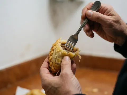 Une main adulte utilise une fourchette pour casser une empanada dorée et croustillante, révélant sa garniture de viande juteuse sur un fond de cuisine flou.