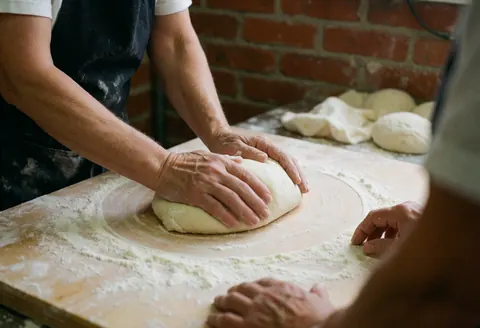 Des mains professionnelles pétrissent de la pâte sur une planche à pain saupoudrée de farine, devant un mur de briques rouges, dans un atelier de cuisine.