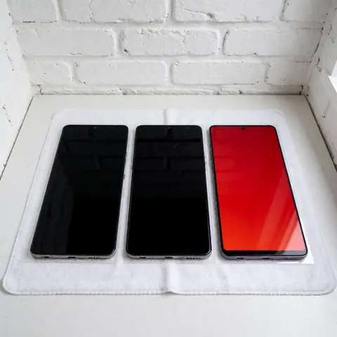Trois téléphones posés sur un tissu blanc, l'un affichant un écran rouge vif, fond de mur en briques blanches, éclairage uniforme, idéal pour la réparation de mobiles.