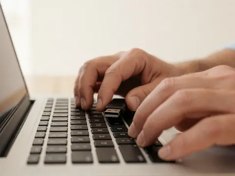 Gros plan sur des mains adultes tapant sur le clavier d'un ordinateur portable avec une clé USB branchée, éclairage doux et fond blanc chaud.