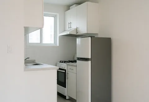 Petite cuisine moderne dans un appartement avec réfrigérateur, cuisinière, armoires blanches et fenêtre laissant entrer la lumière naturelle.