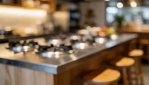 Fond de cuisine moderne avec îlot central, brûleurs en acier inoxydable et tabourets en bois, éclairage chaleureux et ambiance conviviale pour textes.