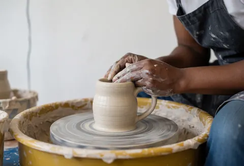 Artiste façonnant une tasse en argile sur un tour de potier professionnel, mains couvertes de terre crue sur fond blanc.