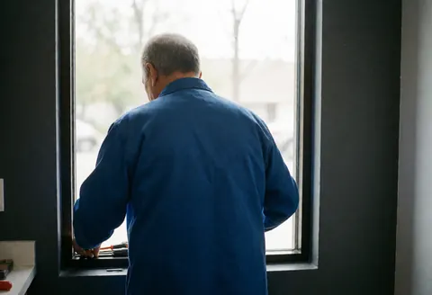 Un technicien en uniforme bleu applique du scellant sur un châssis de fenêtre dans un intérieur aux murs gris, éclairé par la lumière naturelle, mettant en valeur le travail minutieux des mains.