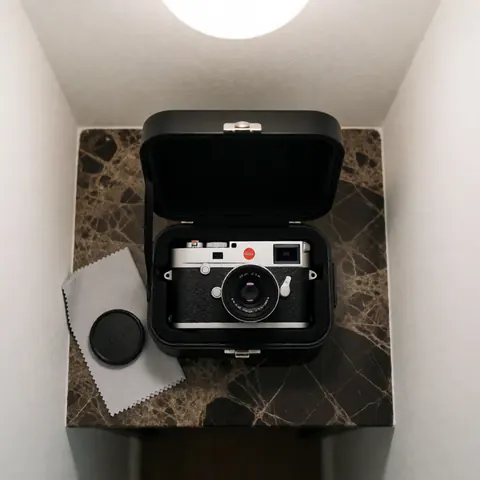 Appareil photo Leica M11 posé dans un étui noir sur une surface en marbre foncé, accompagné d'un chiffon de nettoyage et d'un capot d'objectif, éclairé par une lumière douce venant du dessus.