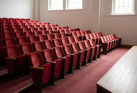 Salle de théâtre avec des sièges rouges et une scène en bois éclairée par la lumière naturelle provenant de fenêtres hautes, créant une ambiance calme et prometteuse.
