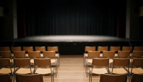 Une scène de théâtre vide avec des chaises en bois disposées face à un fond sombre, éclairage tamisé créant une atmosphère professionnelle pour du texte.
