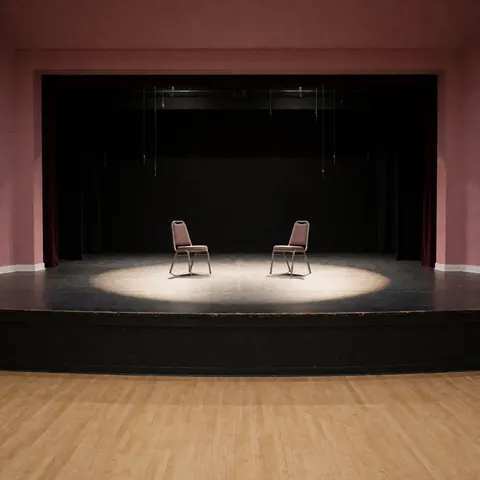 Une scène de théâtre vide avec deux chaises placées sous un faisceau lumineux central, des murs roses et des rideaux noirs en arrière-plan.