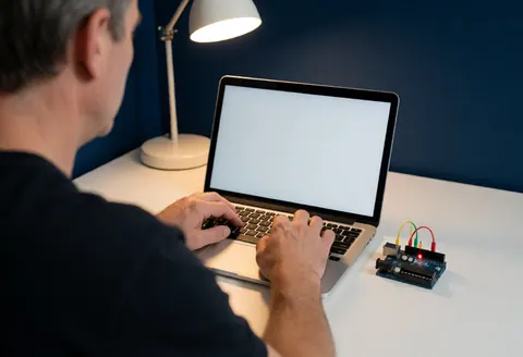Un adulte programme sur un ordinateur portable tandis qu'un Arduino Uno avec des LEDs rouges et vertes clignote sur un bureau blanc sous une lampe de bureau, sur un fond bleu foncé.