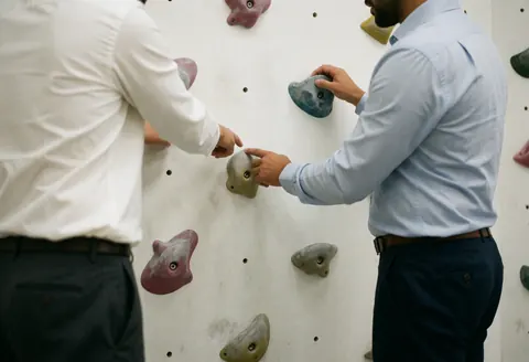 Deux adultes vêtus de chemises et pantalons travaillent ensemble sur un mur d'escalade blanc. L'un indique une prise que l'autre touche, illustrant le travail d'équipe dans un cadre professionnel.