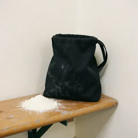 Sac noir de grimpeur posé sur un banc en bois clair, entouré de poussière de craie blanche, murs blancs et éclairage naturel pour une présentation minimaliste.