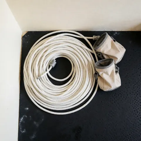 Corde de escalade blanche enroulée en cercle sur un tapis noir perforé, accompagnée de deux sacs de magnésie beige, éclairage uniforme et propre.