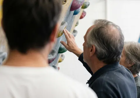 Un instructeur d'escalade pointe vers une prise sur le mur tandis qu'un étudiant est visible de dos au premier plan dans un gymnase lumineux avec des prises colorées.