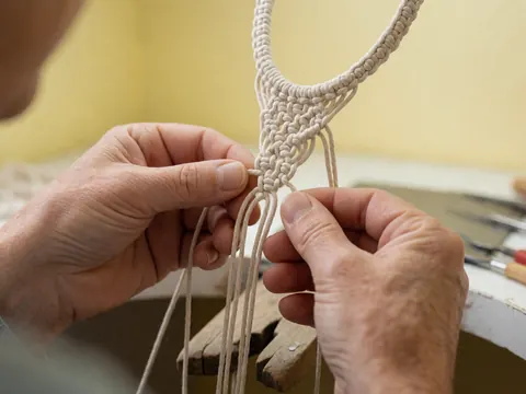 Gros plan sur des mains d'adulte travaillant le macramé avec un cordon blanc sur fond jaune pâle, montrant les détails du tissage en cours.