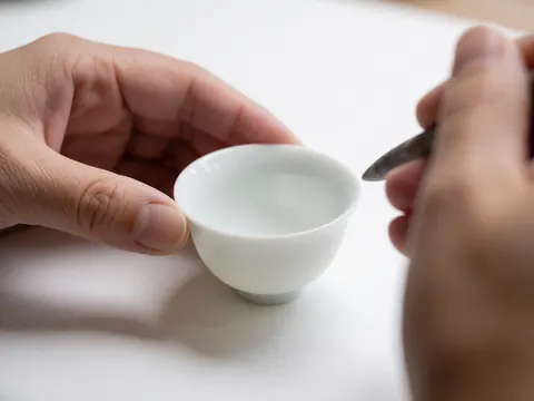 Mains d'un artisan travaillant avec précision une petite coupe en céramique blanche à l'aide d'un outil métallique sur une surface propre.