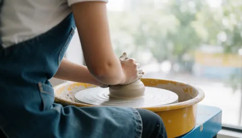 Personne en tablier de jean centrant de l'argile grise sur un tour jaune, éclairée par la lumière naturelle d'une fenêtre dans un atelier calme.