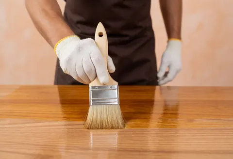 Un professionnel applique du vernis sur une table en bois à l'aide d'un pinceau à poils, portant des gants de protection, sur un fond terracotta.