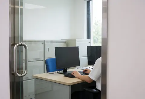 Bureau administratif avec ordinateur, classeurs et fenêtre. Personne travaillant à un bureau. Ambiance professionnelle et propre.