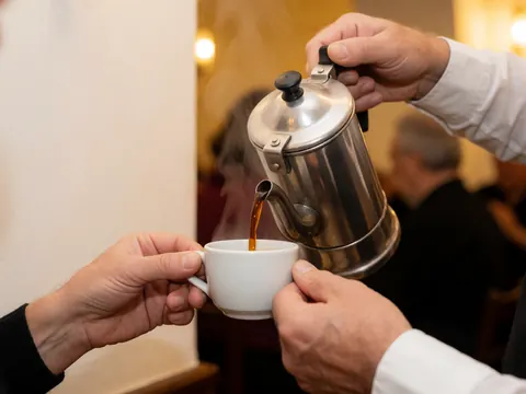 Une main verse du café chaud d'une cafetière métallique dans une tasse blanche sur un fond avec des lumières chaleureuses et des murs clairs.