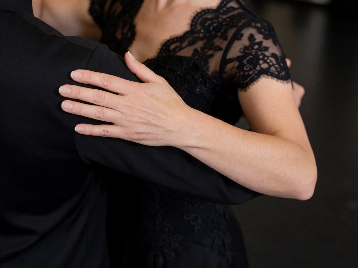 Une main féminine vêtue de dentelle noire saisit l'épaule de son partenaire lors d'une pose de tango, sur un fond sombre et flou soulignant élégance et intimité.