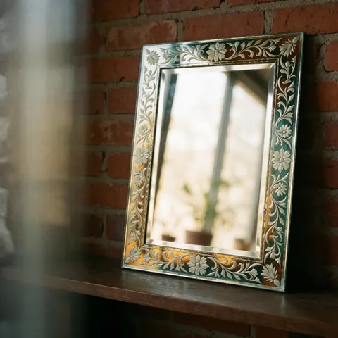 Miroir artisanal au cadre en verre sculpté de fleurs et feuilles, posé sur une étagère en bois devant un mur de briques rouges éclairé par une lumière naturelle douce.