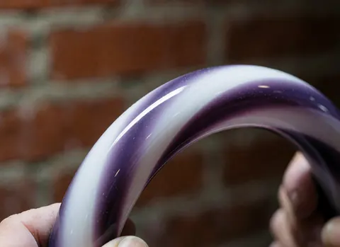 Bague en verre poli avec un dégradé violet et blanc tenue devant un mur de briques rouges mettant en valeur la finition artisanale.