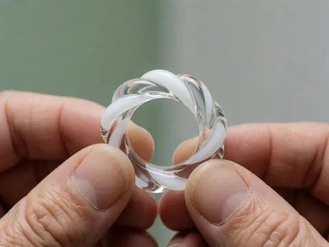 Une bague en verre au motif tressé est soutenue par deux mains sur un fond gris doux avec un mur vert pâle, mettant en valeur les détails de la création artisanale.